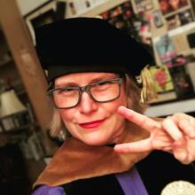Michelle Habeck in a graduation gown and cap, smiling and making a peace sign gesture. She has blonde bob-length hair and is wearing glasses.