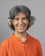 Deborah Beck smiling, with grey hair split down the middle and wearing an orange top, against a grey background.