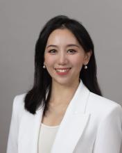 Di Wang, with black hair, is smiling and wearing a white blazer against a grey background.