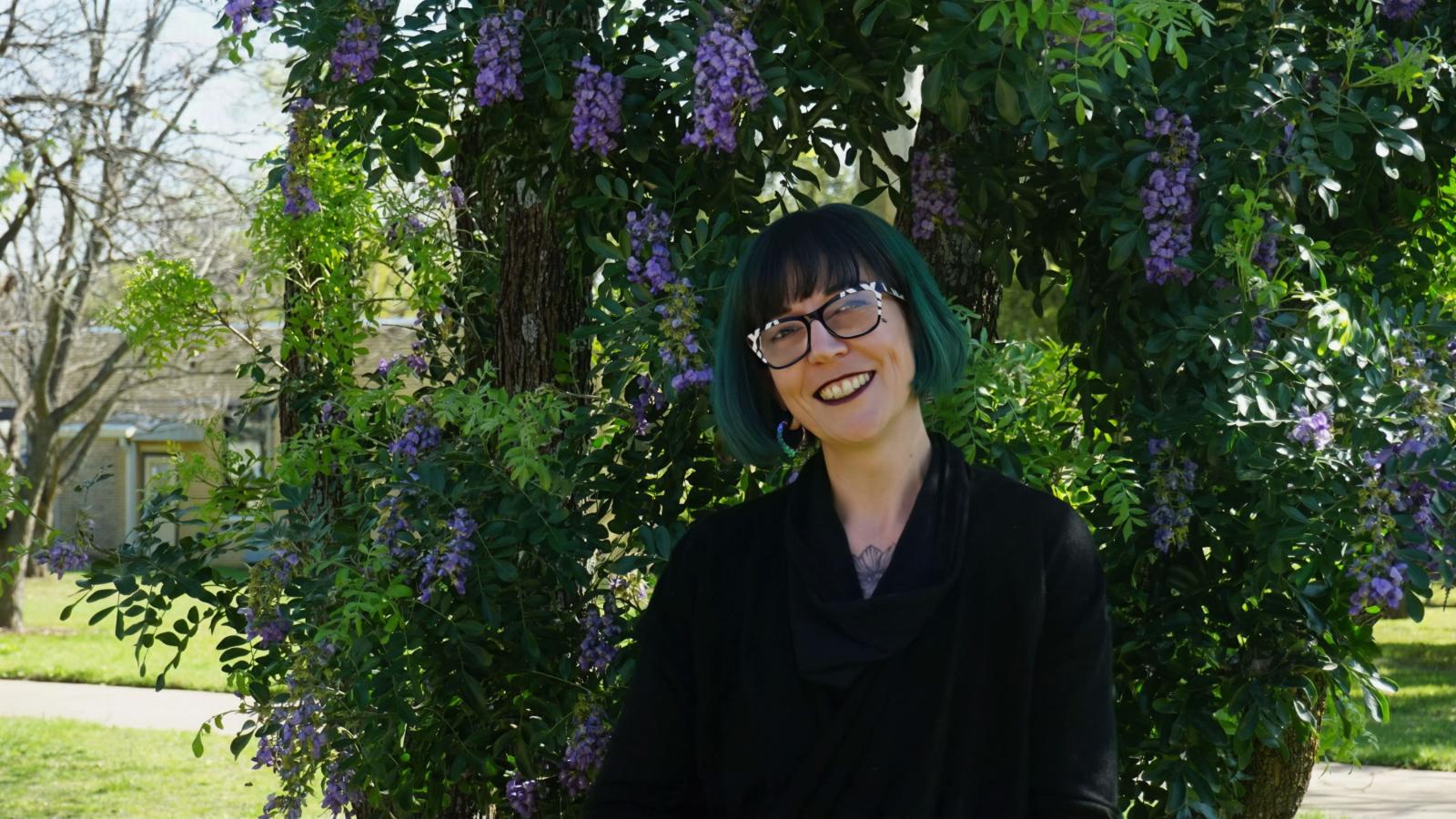 Nick, with black and white glasses and bob-length green hair, smiles while wearing a black top, standing in front of a large tree with purple flowers.