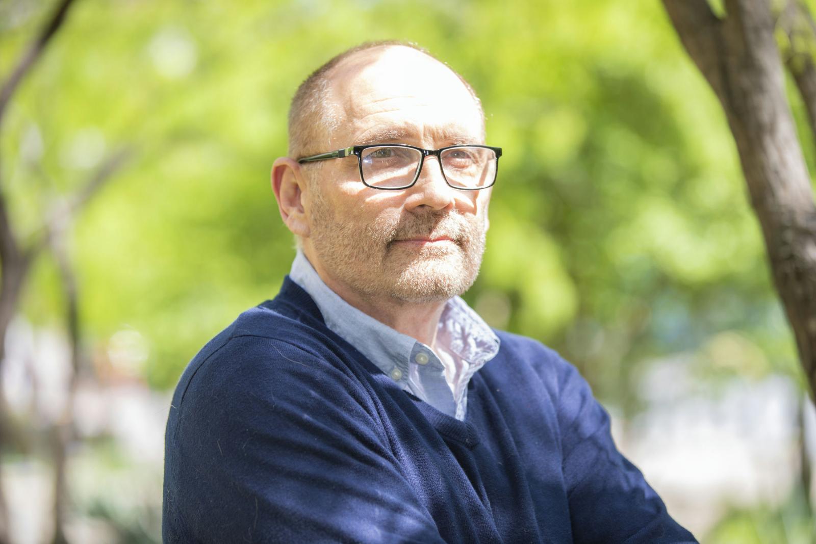 A man wearing glasses and a sweater over a button-up stands in front of trees.