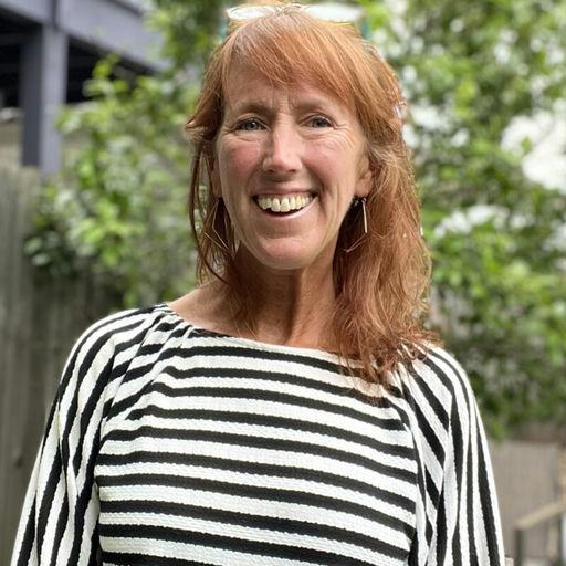 Mary Beer, with red hair, stands in front of a tree, wearing a striped shirt.