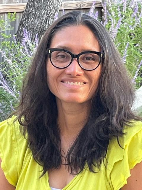 A woman wearing black glasses and a yellow blouse smiles while standing outside.