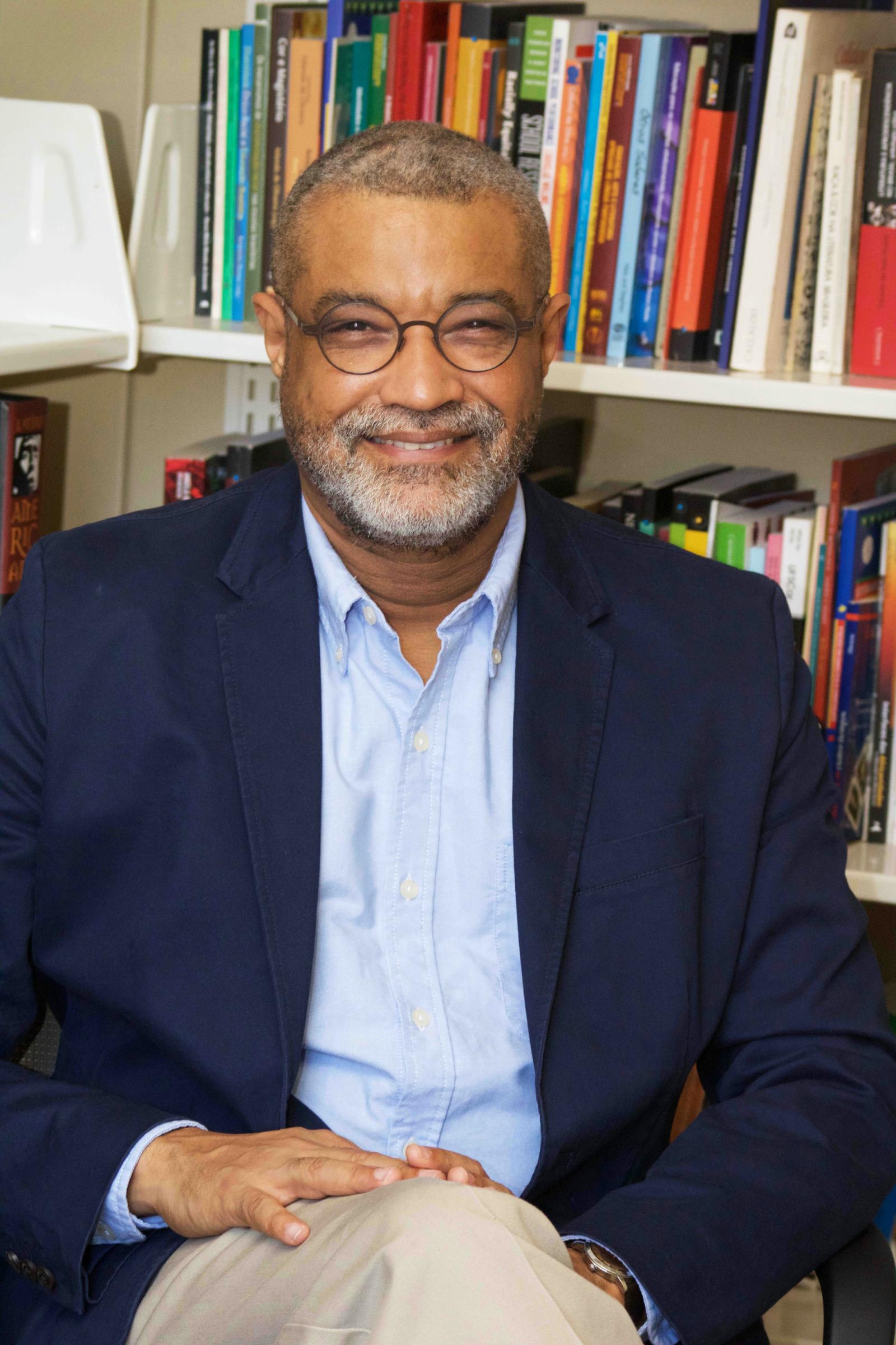 A man with black thin circular glasses and a salt-and-pepper beard smiles while sitting with one leg crossed. He is wearing a blue button-up shirt under a deep dark blue blazer and beige slacks, sitting in front of a bookshelf.