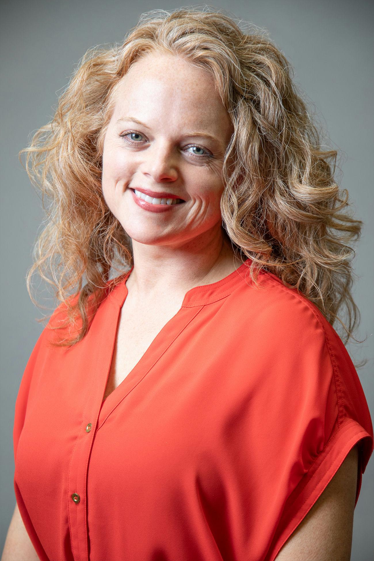 Lara Latimer with curly hair wearing a red shirt in front of a grey background