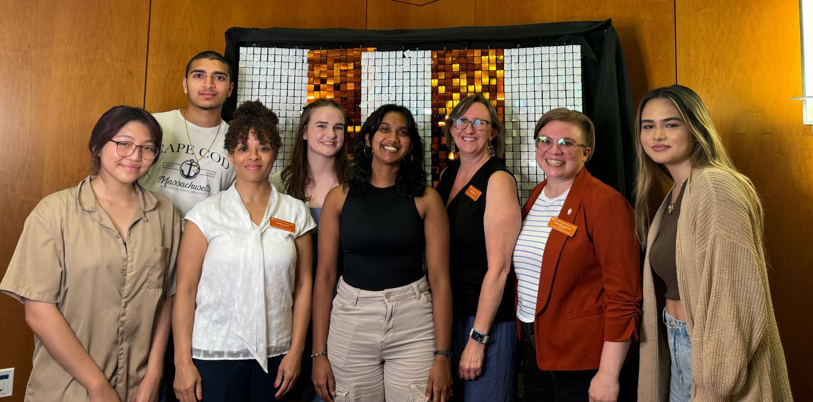 A group of eight people stands indoors in front of a wall with a mosaic pattern. They are smiling and pose together for the photograph.