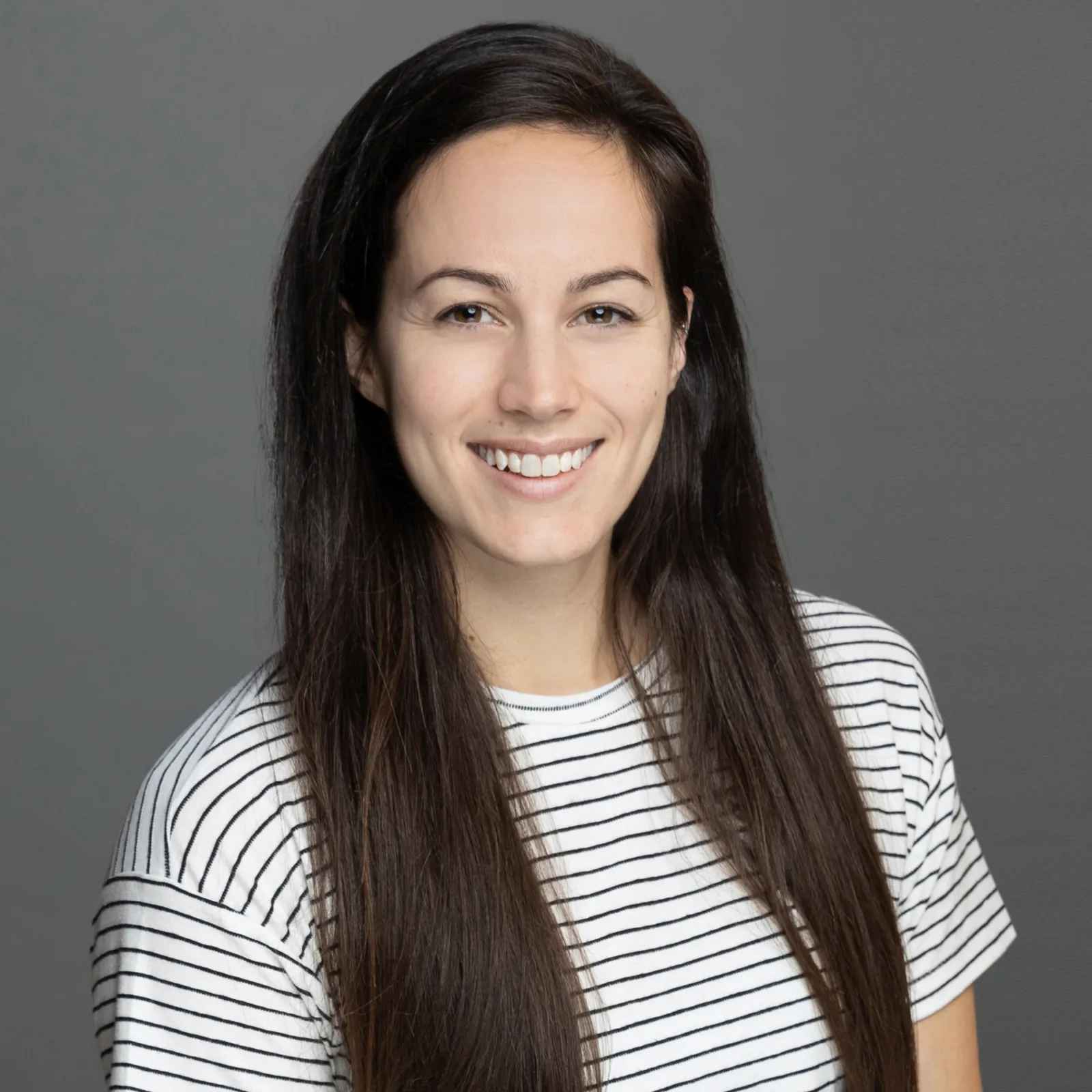 Professional portrait of a woman with long dark hair smiling at the camera against a gray background, wearing a white and black striped top