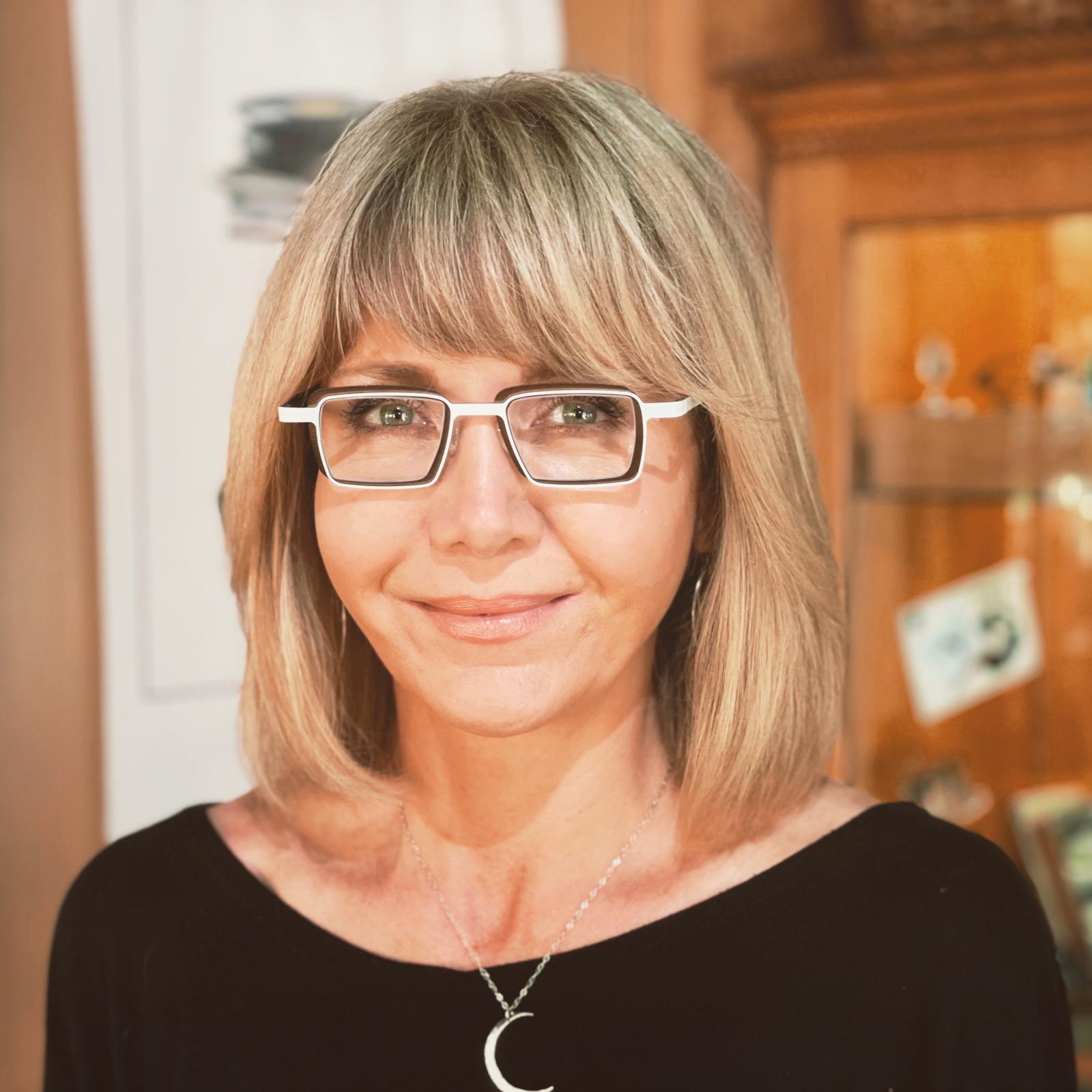 Person with blonde shoulder-length hair with bangs wearing rectangular white glasses, a black top, and a crescent moon pendant necklace, smiling slightly against a warm-toned interior background