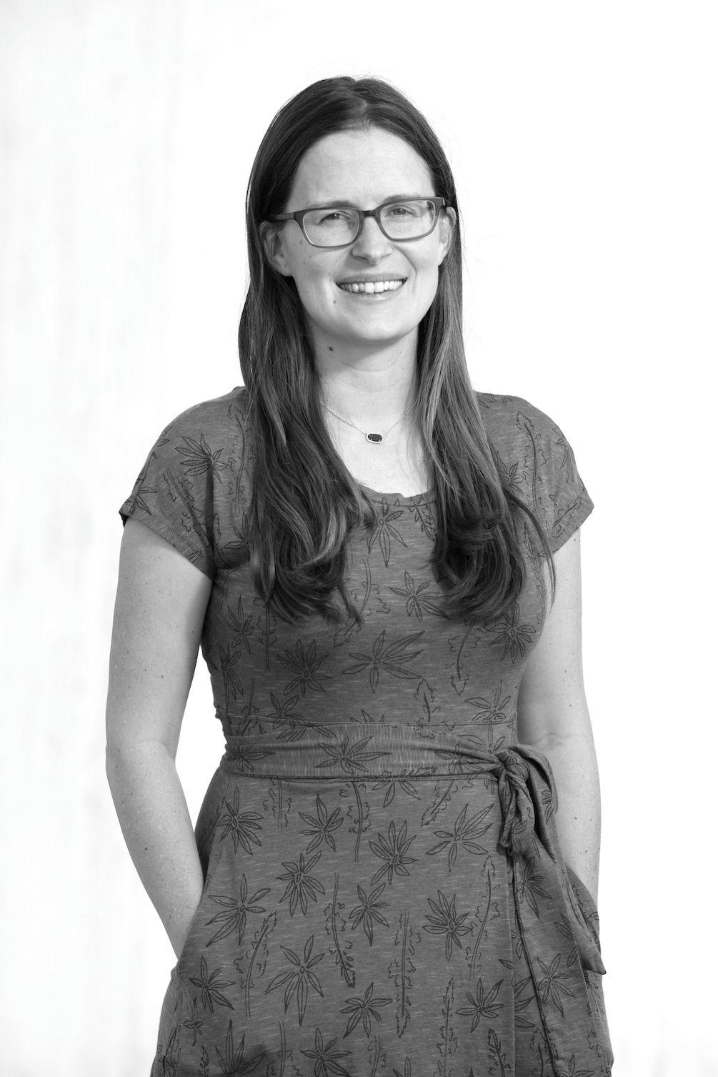 Black and white portrait of a smiling woman with long straight hair and glasses, wearing a patterned dress with hands in pockets.