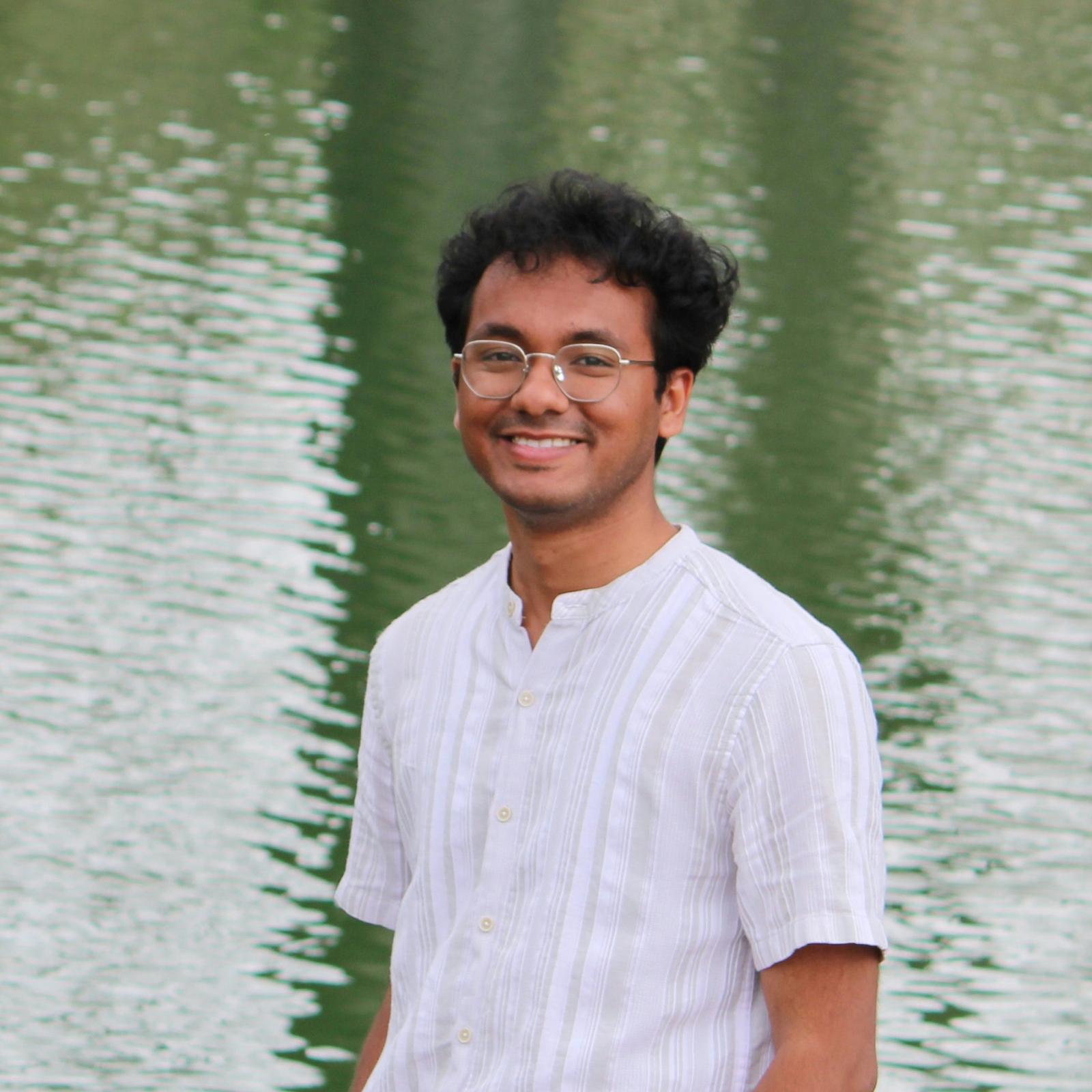 A young man with curly hair and glasses, wearing a white short-sleeve shirt with vertical stripes, smiling while standing in front of a body of water. 