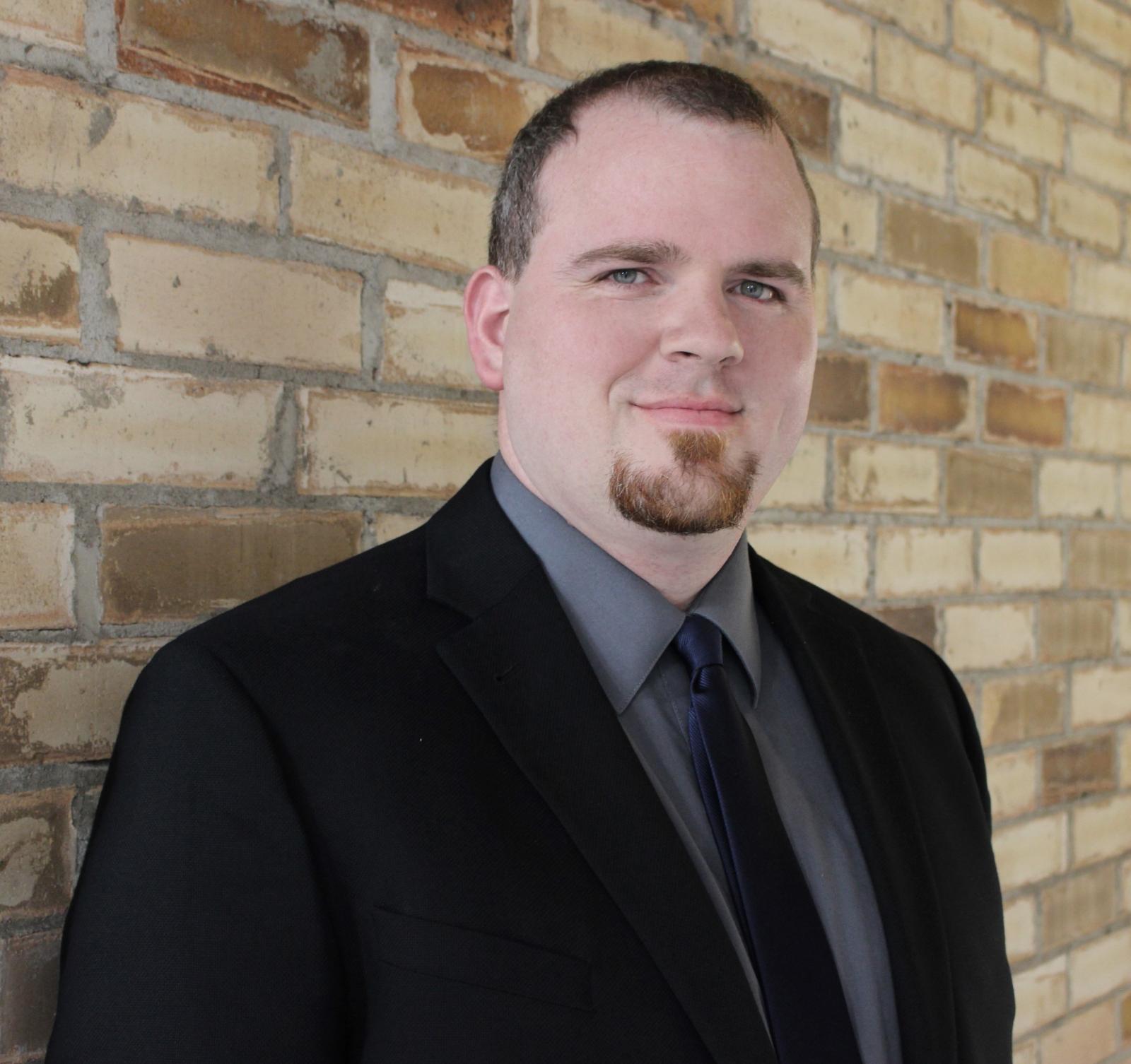 Head and torso of Jonathan Perry, a white man with brown hair and a brown goatee, standing on front of a brick wall.