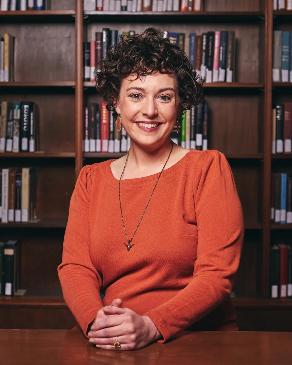 Head and torso of Natalie Czimskey, a white woman with dark brown hair, smiling in front of a bookshelf. person in regalia