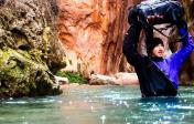 Man crossing a river with water up to his chest & his pack lifted above his head