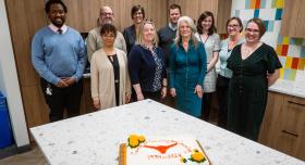 Group of ten people standing behind a kitchen island that has a celebration cake in the foreground