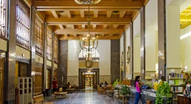 Large rectangular room with high ceilings and chandeliers and various individuals sitting and standing in the distance