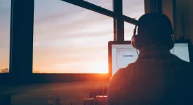 Man at desk with headset in front of computer with sunrise in window 