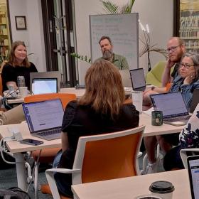 A group of people sit around a table in a library, each with a laptop open in front of them. They appear to be engaged in a discussion or meeting. 