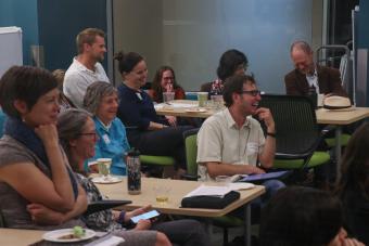 A group of adults are sitting at various tables, smiling, watching an off-screen presentation.