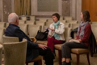 Three adults sitting in a circle, having a discussion, and all three are holding cups of coffee. One woman with short dark hair is gesturing with her hand and speaking, while another woman with long dark hair and a man with grey hair listen.