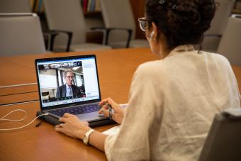 Photo looking over the shoulder of an olive-skinned person with curly dark hair pulled up into a bun. In front of her is a laptop computer, and on the screen a white man with gray hair is giving a speech via videoconferencing.