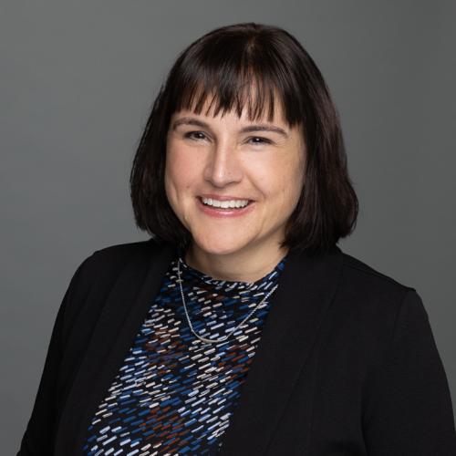 A headshot of Jen Moon, a white woman with dark brown hair, smiling in a black blazer and multicolored shirt.