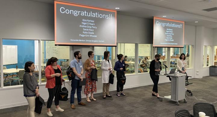 Graduate Student presenters standing in a horizonatl line under a projection screen while they receive certificates.