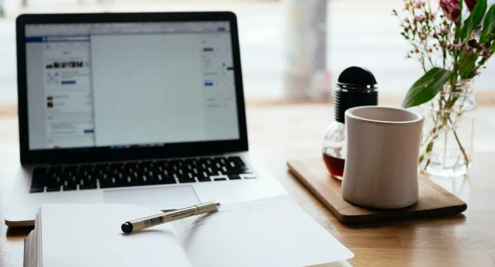 Laptop on a desk with a notebook open in front of it. A pen sits on top of the notebook. A cup of coffee sits to the right of the laptop. This image sourced from Unsplash courtesy of Nick Morrison.