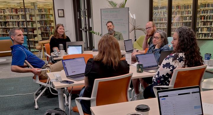 A group of people sit around a table in a library, each with a laptop open in front of them. They appear to be engaged in a discussion or meeting. 