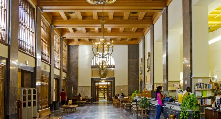 Large rectangular room with high ceilings and chandeliers and various individuals sitting and standing in the distance