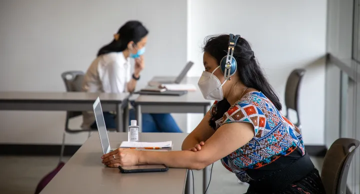 Two individuals masked and working on laptops