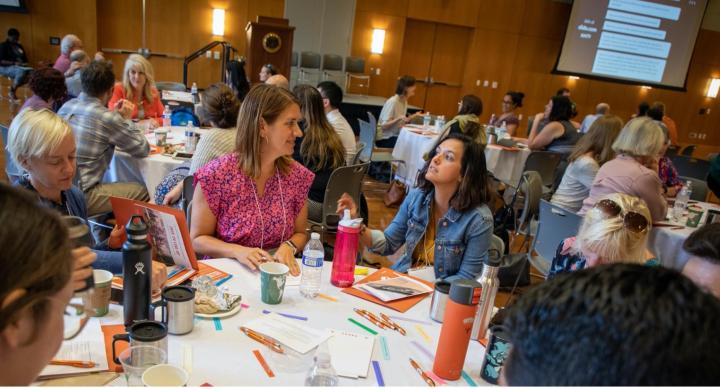 PTF Katie Tackett facilitating a discussion during a New Faculty Symposium 2019 session. Round table with eight people having a discussion. They are generally smiling and looking happy.