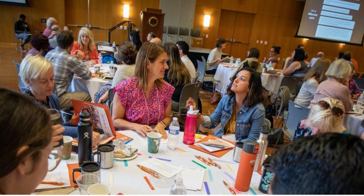 PTF Katie Tackett facilitates a discussion during a New Faculty Symposium 2019 session. Round table with eight people having a discussion. They are generally smiling and looking happy.