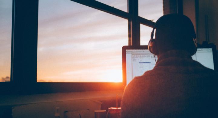 Man at desk with headset in front of computer with sunrise in window 