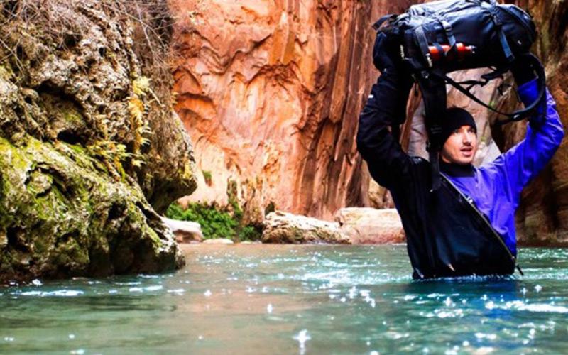 Man crossing a river with water up to his chest & his pack lifted above his head